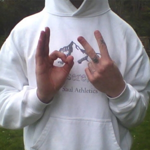 A man wearing a white hooded sweatshirt with a mountain logo makes two hand gestures ('b' and '2'/peace sign) while standing in a grassy outdoor area