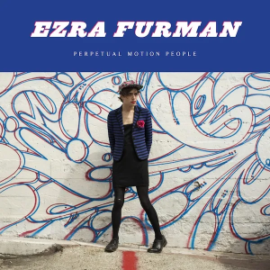 Ezra Furman standing in front of a white wall painted with abstract blue and red line art.