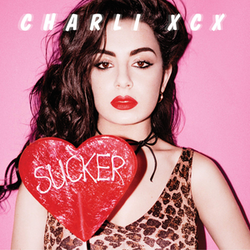 Charli xcx wearing red lipstick and a leopard-print top against a bright pink background, holding a large heart-shaped red lollipop with the word 'SUCKER' written on it in white
